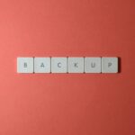 Close-up of keyboard keys spelling 'BACKUP' placed on a coral-colored surface.